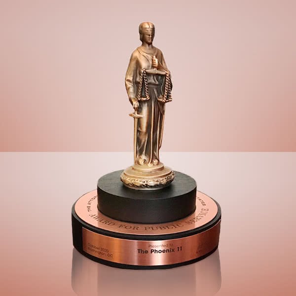 The Phoenix 11’s award for Meritorious Public Service: a cast figure of Lady Justice in robes wearing a blindfold, holding scales in one hand and a sword in the other with its point resting beside her feet, standing on top of a disc describing the award’s purpose.